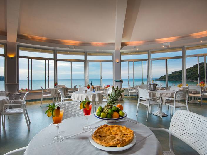 Bright restaurant with sea views, featuring colorful cocktails, fresh fruit, and a pie on elegant white tables.