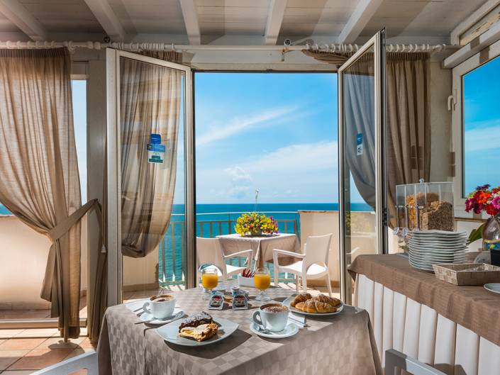 A bright dining area with ocean views, featuring a table set with pastries, coffee, and fresh flowers.