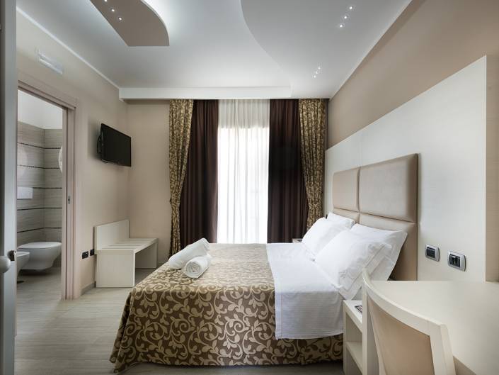 Modern hotel room with a large bed, soft curtains, a TV, and an ensuite bathroom in neutral colors in Cortona, Arezzo